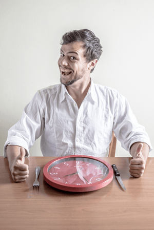 Man smiling at table with red wall clock, fork, and knife, illustrating meal timing